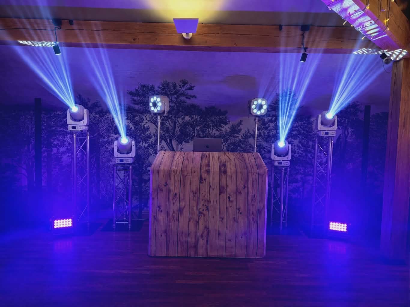Rustic wooden DJ booth in front of a forest mural, lit by four moving-head lights